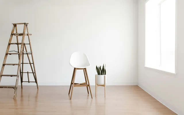 Una habitación clara y vacía, con una escalera de tijera de madera, una silla sola y una planta sobre suelo de madera.