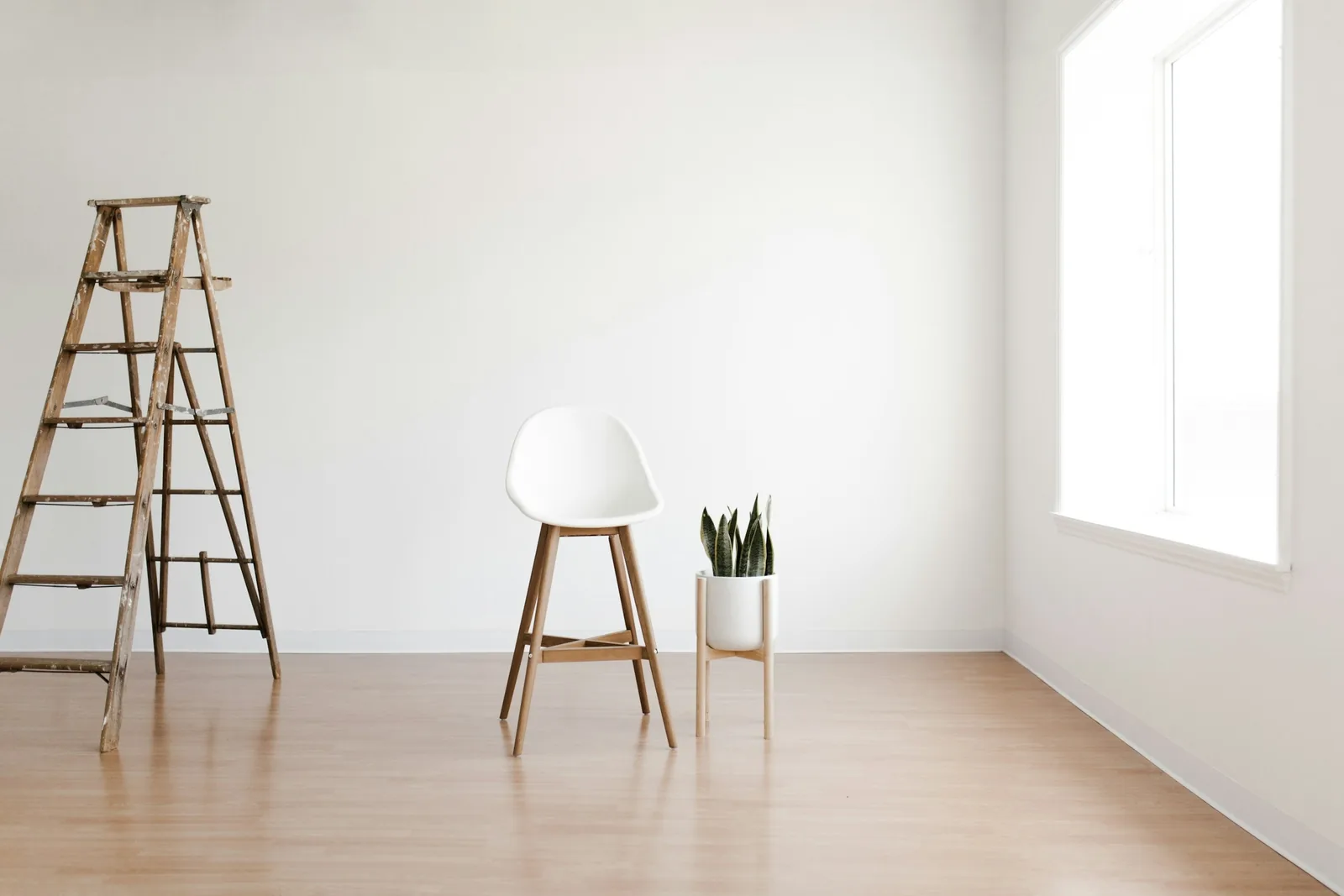 Una habitación clara y vacía, con una escalera de tijera de madera, una silla sola y una planta sobre suelo de madera.