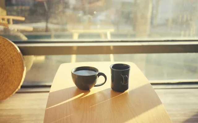 Dos tazas sobre una mesa de madera junto a una ventana, con la luz dorada de la tarde entrando de frente.